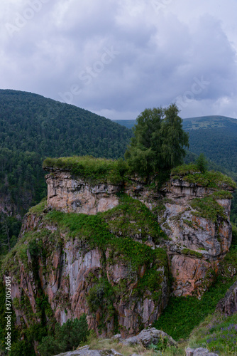 the roadbed is uneven, winding, passes through mountains and cliffs. Clouds are dark, low, with rain. A thick white fog is spreading along the road.