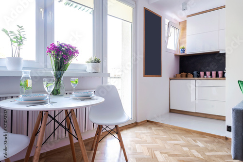 White interior of dining room with window, table, chair, dinnerware and flower. Wooden floor and small kitchen in cozy indoor. Scandinavian design.