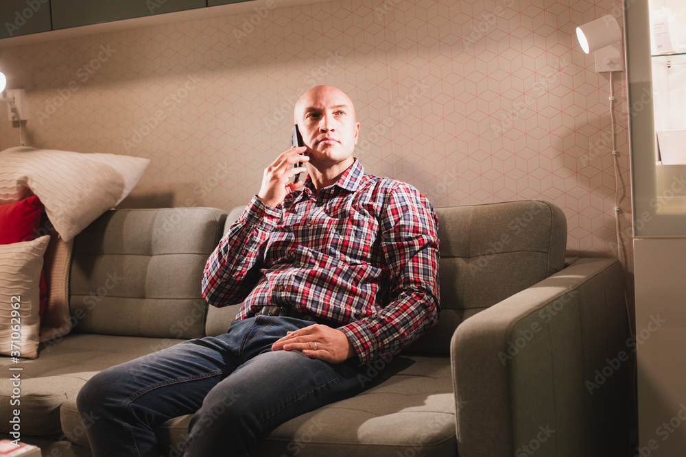 An adult bald man, dressed in a plaid shirt and jeans, is sitting on the couch and talking on a smartphone. Positive communication.