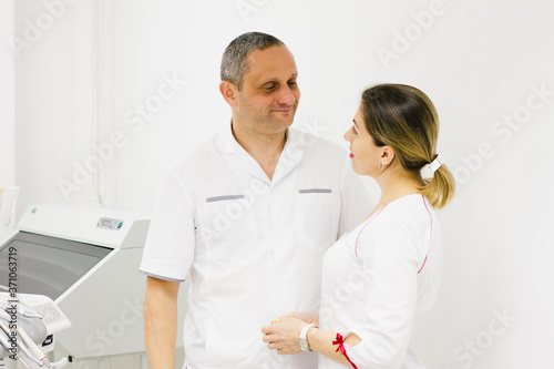 Two professional cheerful doctors  a senior man and a young woman with bright lipstick  dressed in white coats against the background of medical equipment. Health concept.