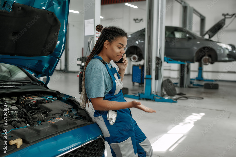 Reliable car doctor. Young african american woman, professional female  mechanic talking on phone, leaning on a car with open hood at auto repair  shop Photos | Adobe Stock