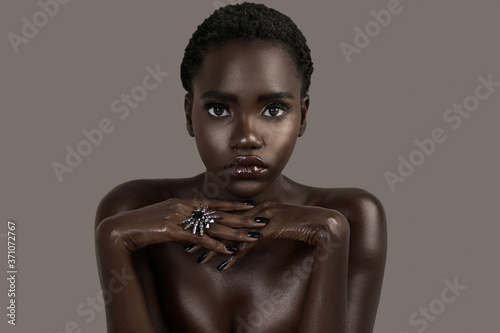 A portrait of a serene young black female with short black hair, moist lips, perfectly black manicured nails with silver jewelry looking straight at the camera.