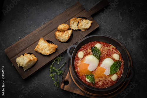 hearty English breakfast - poached eggs, with tomatoes and sauce in a pan on a wooden board 
