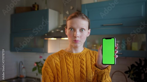 Beautiful young woman looks surprise at phone, shows green screen looks at camera shows gesture cool on kitchen