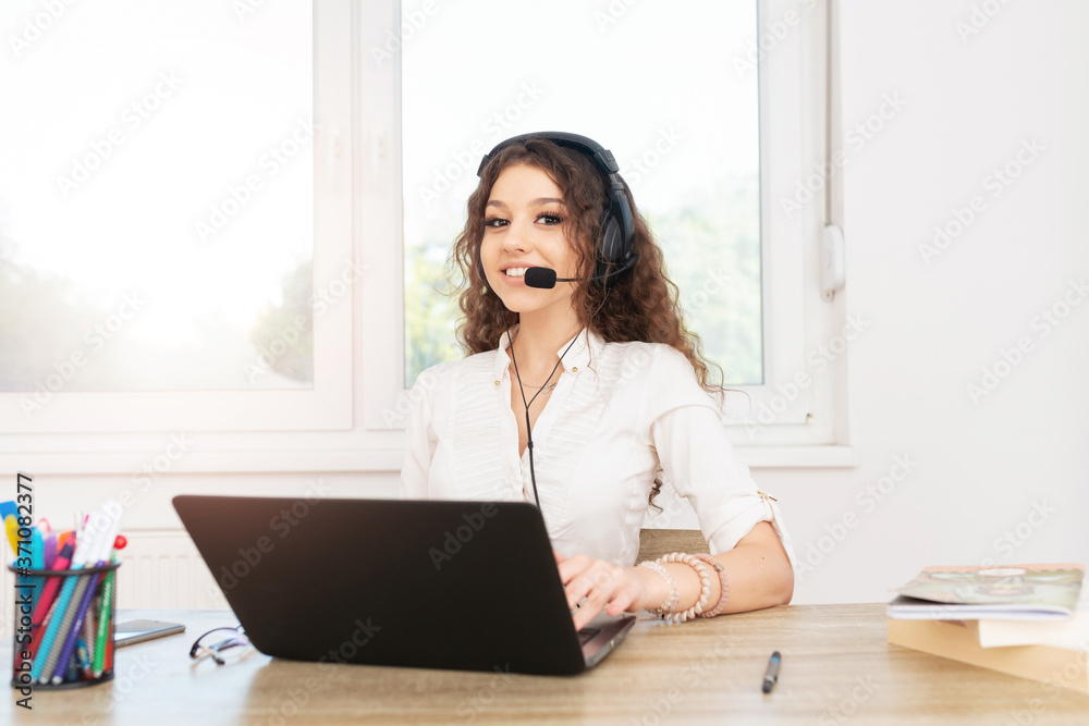 Professional young woman with curly brown hair sitting in office by the window and using laptop and headphones. Professional young woman with curly brown hair sitting in office by the window and using laptop and headphones.