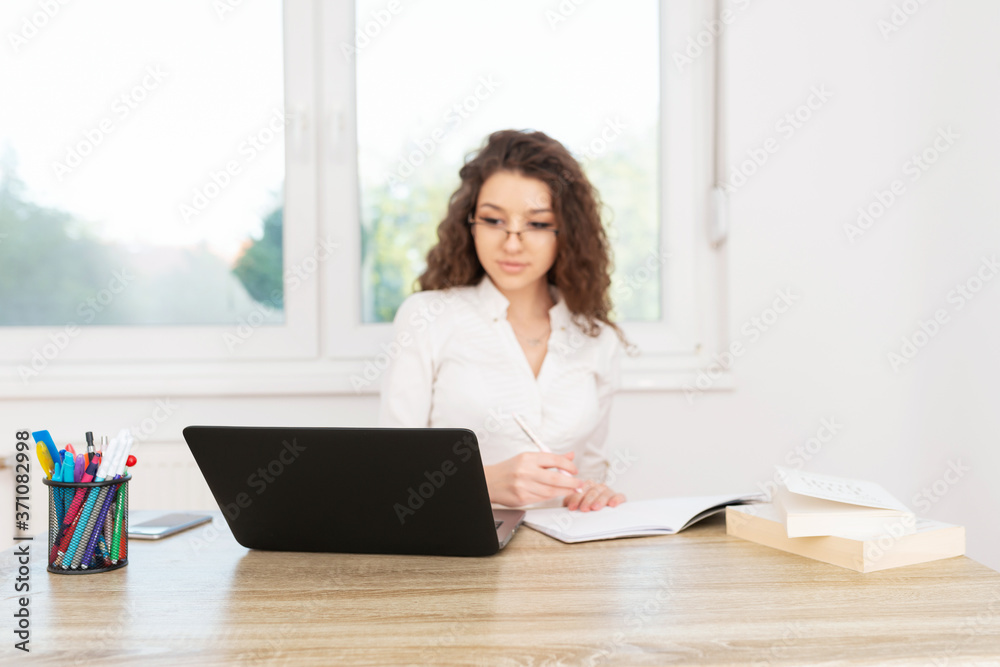 Corporate female with curly hair, sitting in office by the window, wearing glasses and looking at laptop. Corporate female with curly hair, sitting in office by the window, wearing glasses and looking at laptop.
