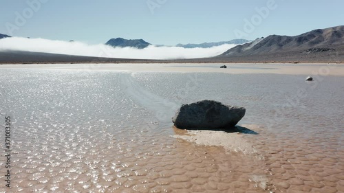 Close up of black rock on the wet cracked surface of Death Valley desert. Epic race track playa park. Cinematic 4K aerial of moving stones on sunny morning, with mountains and clouds on background