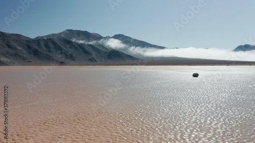Drone flying around black rock on wet cracked surface of Death Valley desert. Epic race track playa park. Cinematic 4K aerial of moving stones on sunny morning, with mountains and clouds on background
