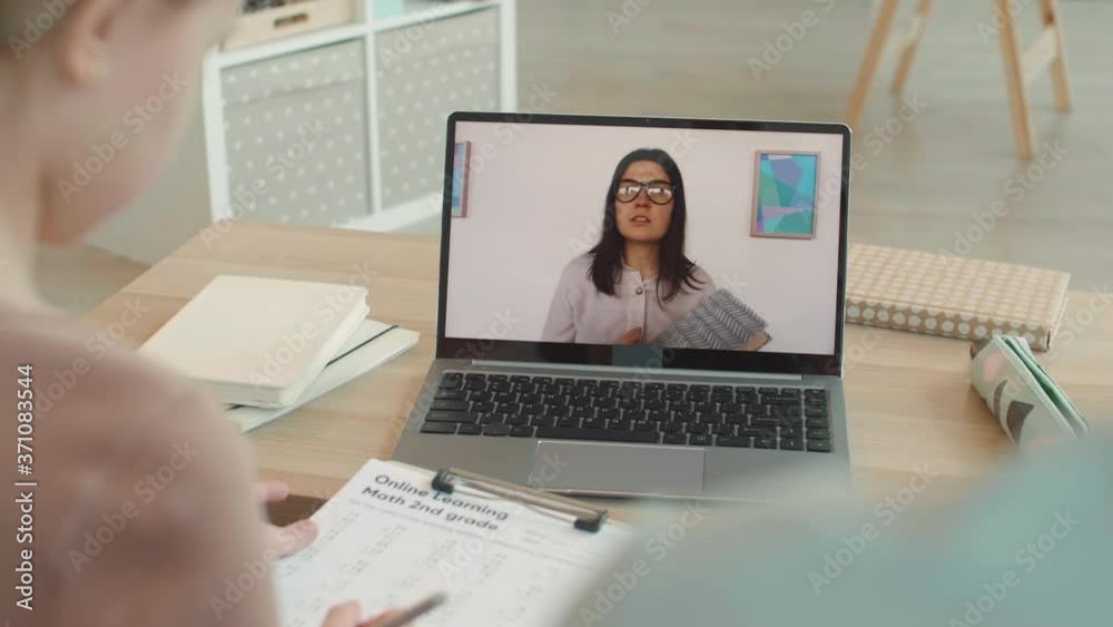 Rear view of Caucasian schoolgirl is sitting at desktop at home and listening to young female teacher explaining math task by video link Rear view of Caucasian schoolgirl is sitting at desktop at home and listening to young female teacher explaining math task by video link
