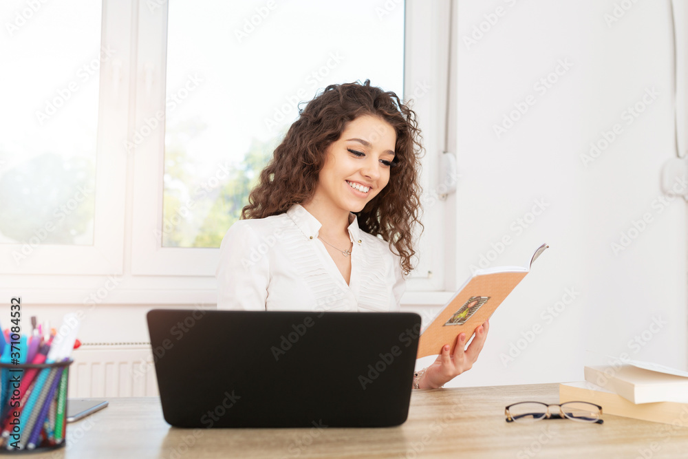Elegant businesswoman in white shirt sitting at work desk with laptop, reading a book and smiling. Elegant businesswoman in white shirt sitting at work desk with laptop, reading a book and smiling.