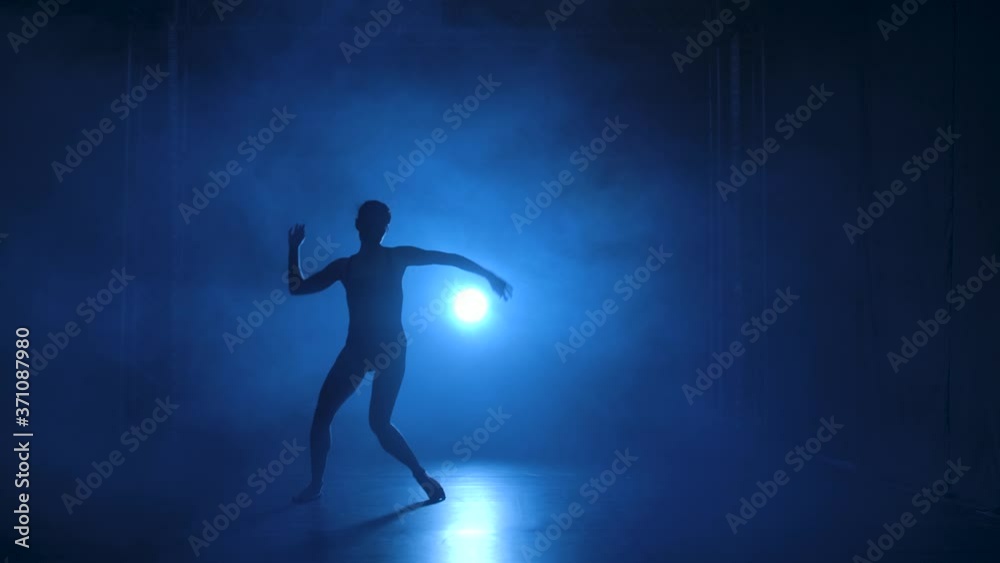 Silhouette a professional ballerina in black bodysuit dancing flexible in darkness under a theatrical blue spotlights of studio. Slow motion.