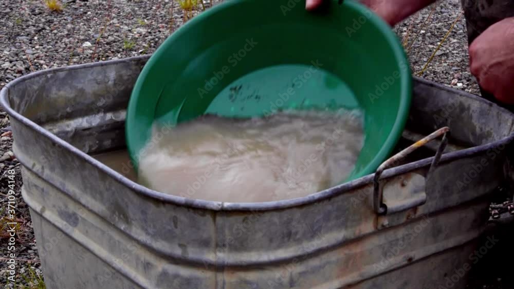 Gold panning black fine grunge, silt, sand, clay, and gravel alluvial deposits in green trap pan, wetting, soaking, agitating and washing in brown tin bucket of murky water, static close up portrait Gold panning black fine grunge, silt, sand, clay, and gravel alluvial deposits in green trap pan, wetting, soaking, agitating and washing in brown tin bucket of murky water, static close up portrait