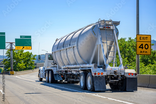 Big rig white day cab semi truck tractor transporting cargo in closed bulk semi trailer running on the highway exit intersection