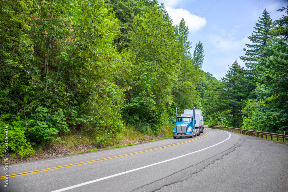 Blue day cab big rig semi truck with vertical pipes transporting covered cargo on flat bed semi trailer running on the winding forest road in Columbia Gorge Area Blue day cab big rig semi truck with vertical pipes transporting covered cargo on flat bed semi trailer running on the winding forest road in Columbia Gorge Area