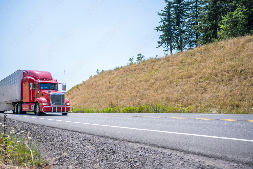 Powerful red big rig diesel semi truck carry cargo in refrigerator semi trailer running on the summer winding road with hill and trees
