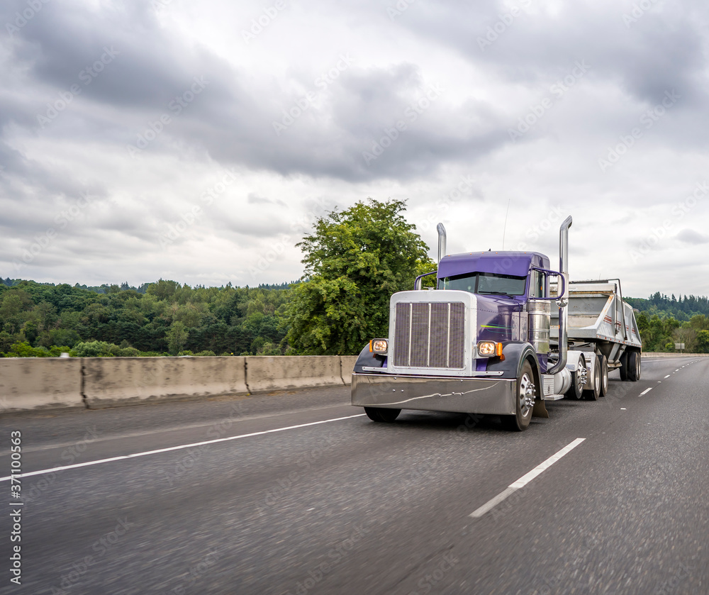Stylish purple big rig classic semi truck with vertical pipes transporting cargo in tipper semi trailer running on the wide straight highway road