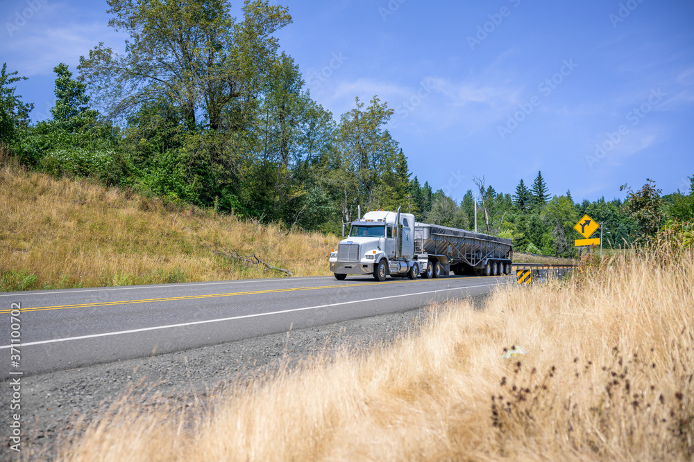 White classic big rig semi truck transporting cargo in covered bulk semi trailer running on the road with hills on the sides in summer sunny day