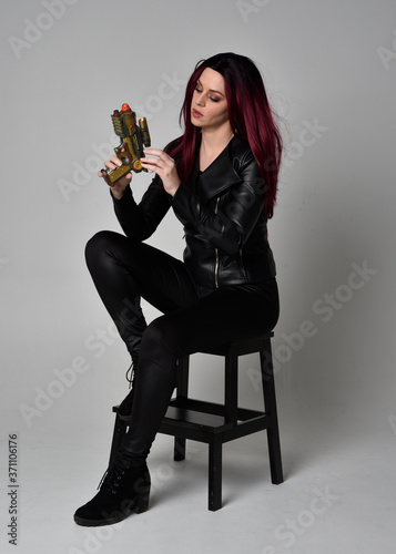 Full length portrait of girl with red hair wearing black leather jacket, pants and boots. Sitting pose on a chair holding a gun, isolated against a grey studio background.