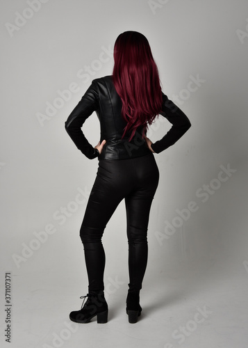 Full length portrait of  girl with red hair wearing black leather jacket, pants and boots. Standing pose facing away from the camera, isolated against a grey studio background.