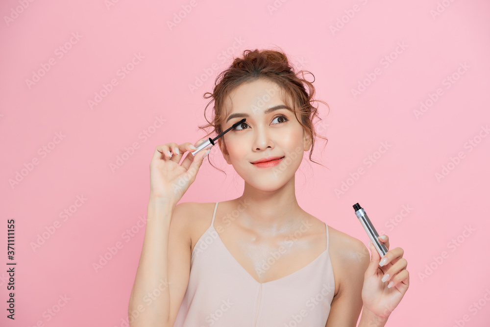 Young beautiful Asian woman applying black mascara on eyelashes with makeup brush over pink background. Young beautiful Asian woman applying black mascara on eyelashes with makeup brush over pink background.