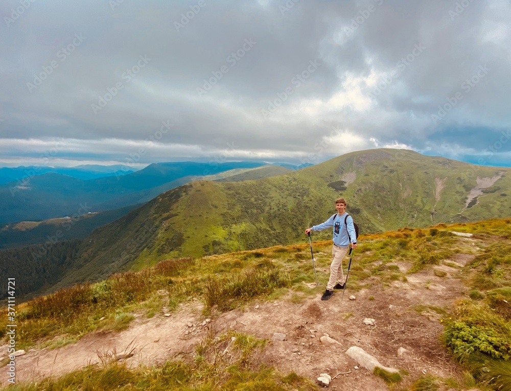 The young blonde guy is in the mountains. Blue sky and beautiful nature. Fit man climb. Green trees and green grass