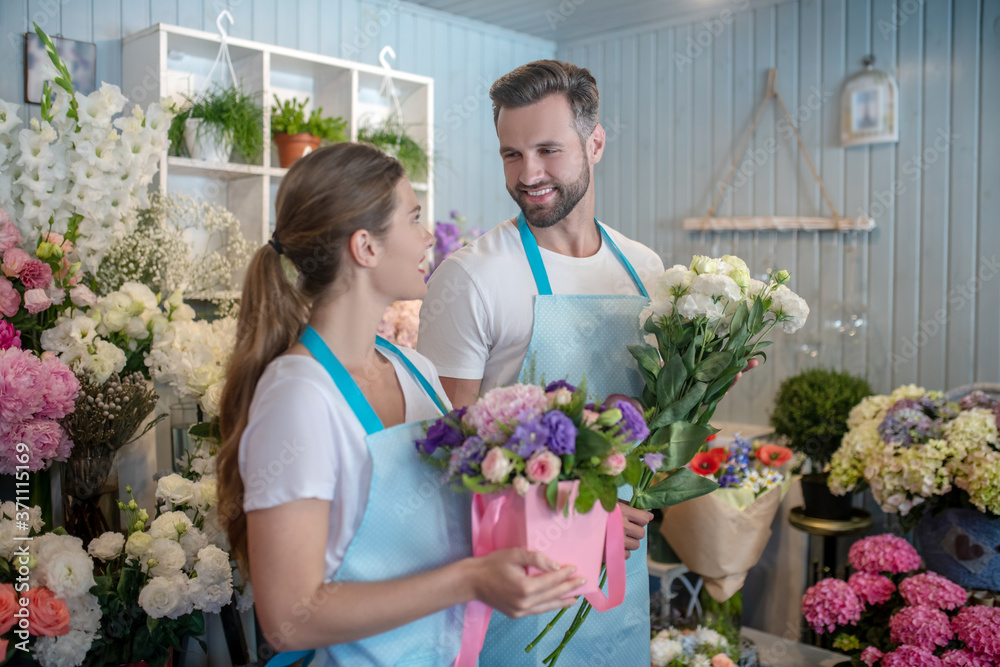 Bearded male and brown-haired female in aprons holding flowers, looking at each other, smiling