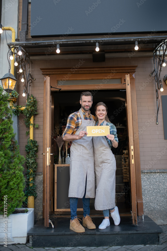 Young waitress and bearded waiter standing at the entrance, holding yellow open sign, smiling