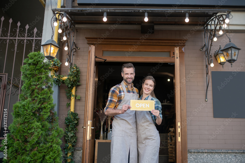 Young waitress and bearded waiter in grey aprons standing outside, holding yellow open sign