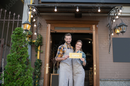 Young waitress and bearded waiter in grey aprons standing outside  holding yellow open sign