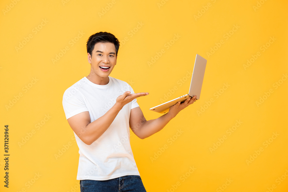 Portrait of smiling young handsome Asian man presenting laptop while smiling and looking at camera in isolated studio yellow background Portrait of smiling young handsome Asian man presenting laptop while smiling and looking at camera in isolated studio yellow background