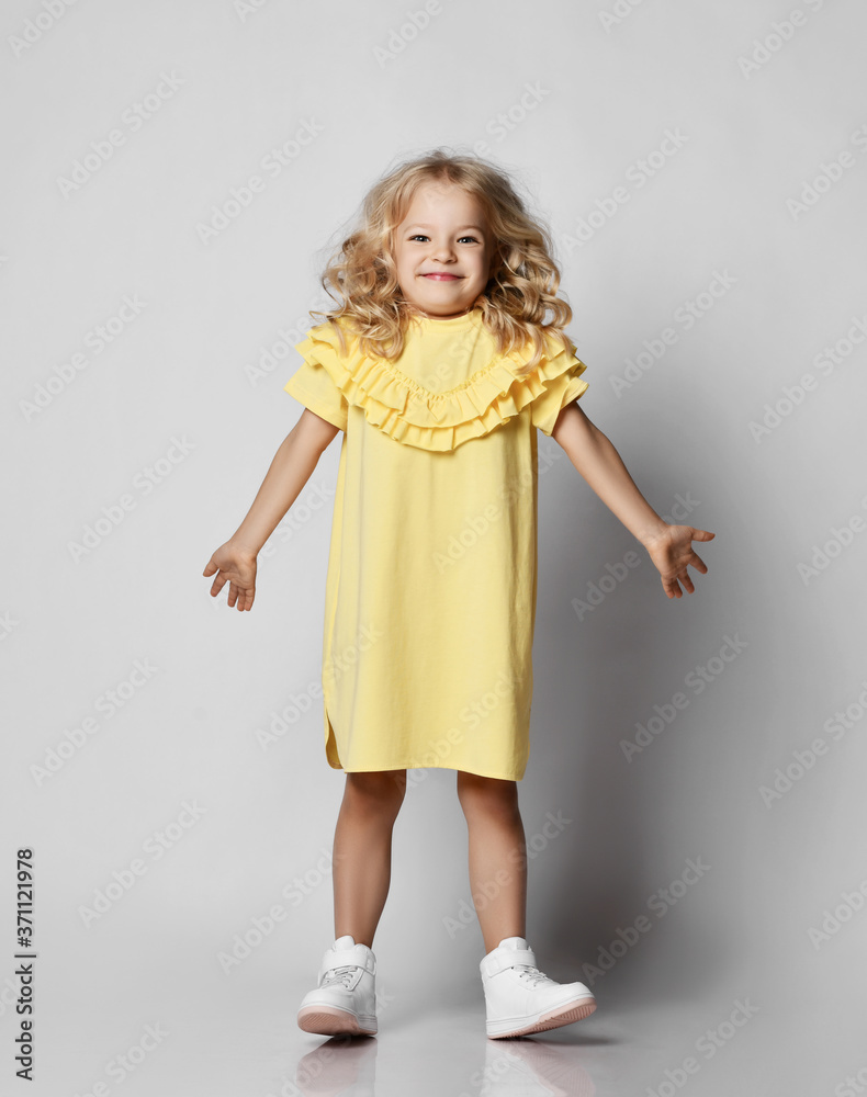 Frolic playful blonde curly kid girl in yellow dress and white sneakers is having fun, funny posing, playing Frolic playful blonde curly kid girl in yellow dress and white sneakers is having fun, funny posing, playing