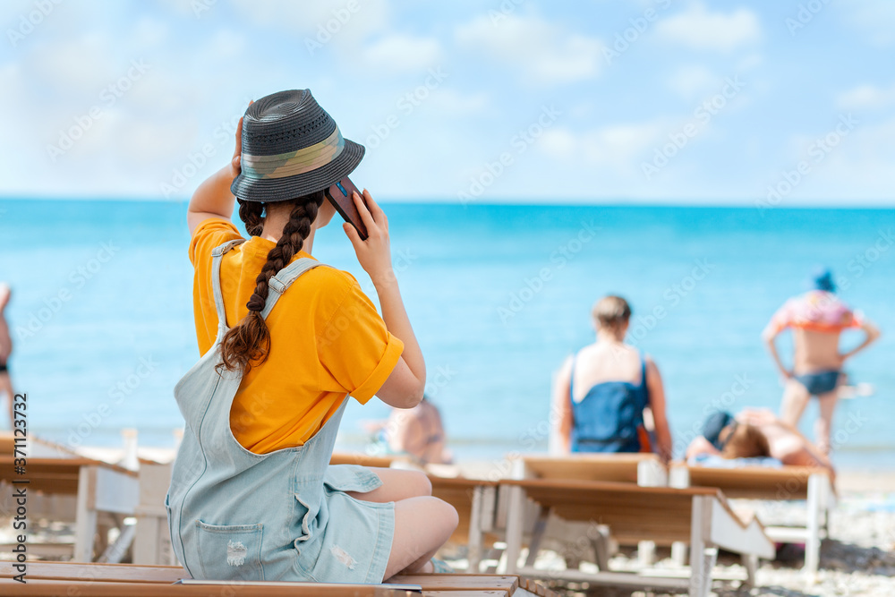 A woman in a straw hat is sitting on a sunbed and making a phone call. Rear view. Sea and sky in the background. Copy space