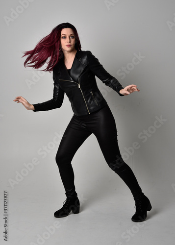 Full length portrait of girl with red hair wearing black leather jacket, pants and boots. Standing pose, isolated against a grey studio background.