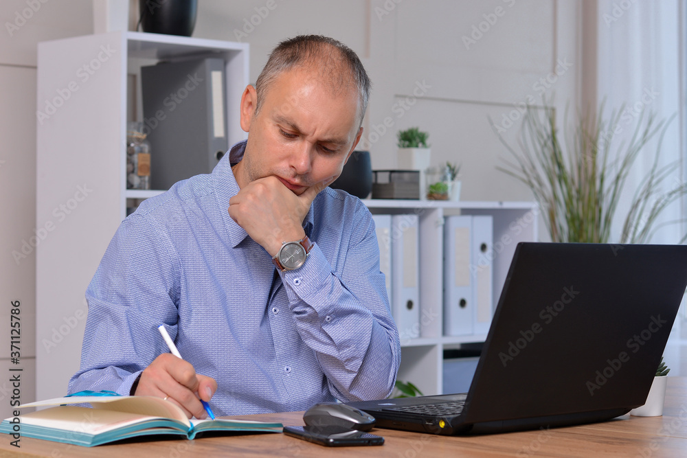 A pensive businessman working at a laptop writes about his plans and goals in his notepad A pensive businessman working at a laptop writes about his plans and goals in his notepad