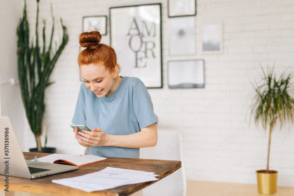 Portrait of happy cheerful redhead young business woman using mobile phone while sitting at the desk with laptop in light cozy room with modern interior at home office. Portrait of happy cheerful redhead young business woman using mobile phone while sitting at the desk with laptop in light cozy room with modern interior at home office.