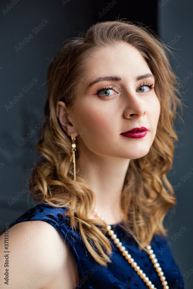 Naklejka premium A beautiful girl in a blue velvet dress and pearl beads near the window.