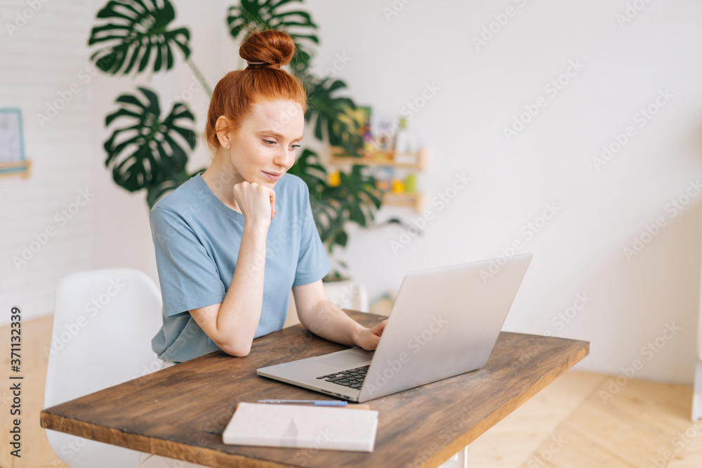 Cute redhead young businesswoman typing on laptop computer at the desk in light cozy room at the home office. Concept of distance job on Internet in self isolation. Cute redhead young businesswoman typing on laptop computer at the desk in light cozy room at the home office. Concept of distance job on Internet in self isolation.