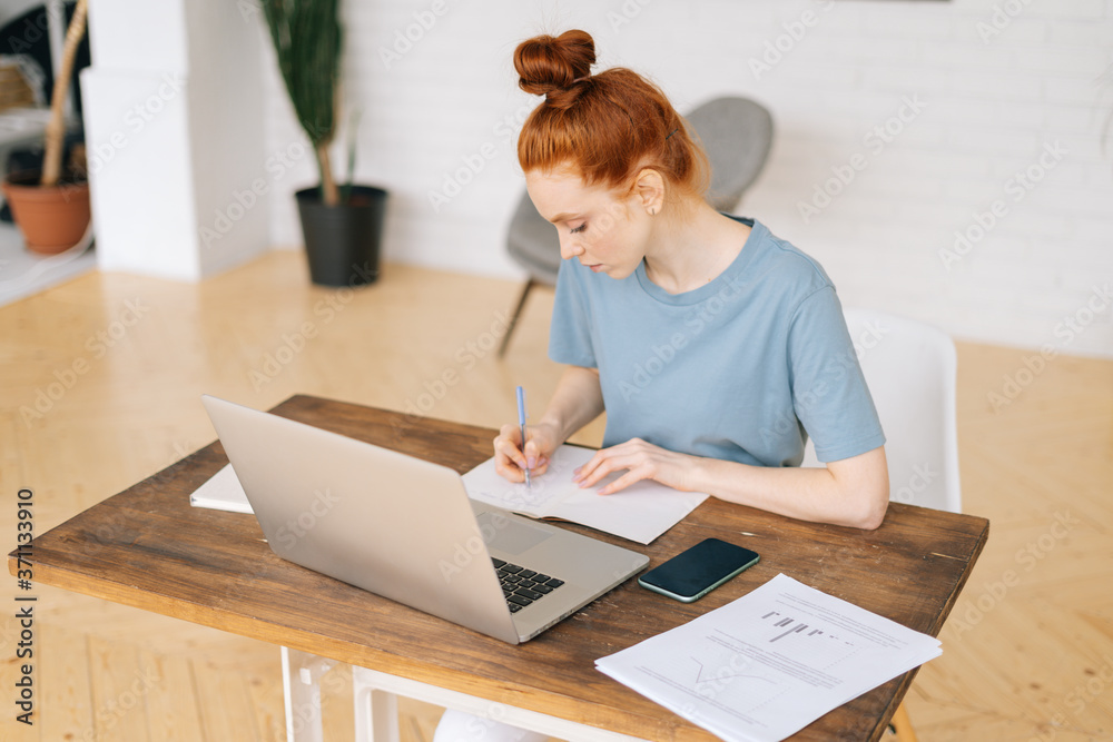 Creative redhead young woman designer is writing in documents, working on laptop while sitting at the desk in light cozy living room at home office. Concept of remote working. Creative redhead young woman designer is writing in documents, working on laptop while sitting at the desk in light cozy living room at home office. Concept of remote working.