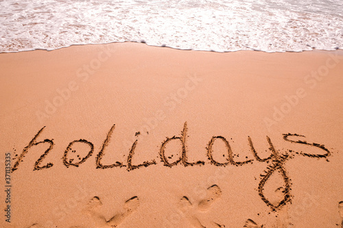 Word Holidays hand written in the sand with a sea wave. Close up sand texture on beach in summer.
