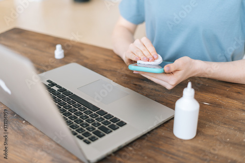 Close-up of hands of unrecognizable woman wiping with antibacterial antiseptic sanitizer spray display screen of cell phone while sitting at the table with laptop at home office.