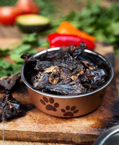 Dried beef liver in the dog vessel among the greenery on the wooden board and some vegetables on the background. Chewing treats for domestic dogs. Natural dental treats for promotion and pampering.