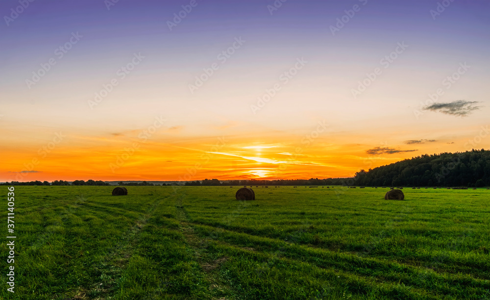 Scenic view at beautiful sunset in green shiny field with hay stacks, bright cloudy sky, country road and golden sun rays with glow, summer valley landscape
