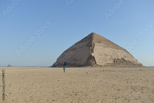 Obraz na płótnie a tourist walks alone through the desert in front of The Bent Pyramid an ancient Egyptian pyramid located at the royal necropolis of Dahshur w salonie