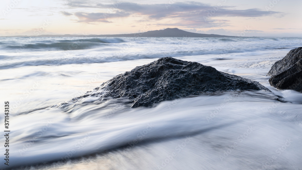 Waves crashing over rocks at sunrise with Rangitoto island in the distance.