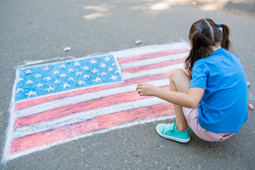 Two cute friends girls drawing American flag with colored chalks on the sidewalk near the house on sunny summer day. Kids painting outside. Creative development of children. Patriotic day concept