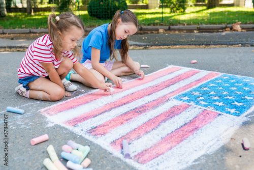 Two cute friends girls drawing American flag with colored chalks on the sidewalk near the house on sunny summer day. Kids painting outside. Creative development of children. Patriotic day concept