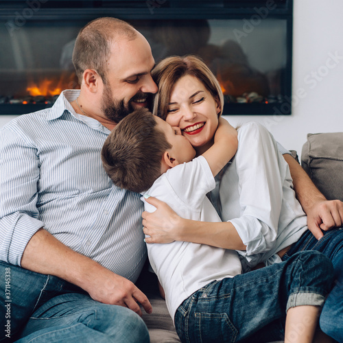 caucasian family is sitting on a gray sofa at home cuddling up and looking at the camera. Father hugs his wife and son