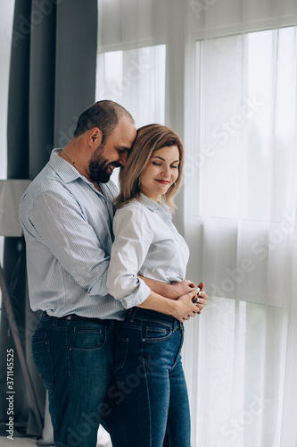 a woman and a man in jeans and shirts stand near a panoramic window and hug gently. Man embracing girlfriend at home © ElenaKyrylova