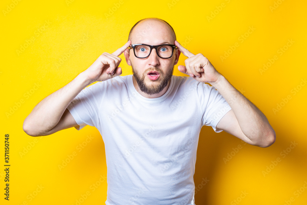 Serious bearded bald young guy with glasses holds fingers at his temple on a yellow background. Serious bearded bald young guy with glasses holds fingers at his temple on a yellow background.