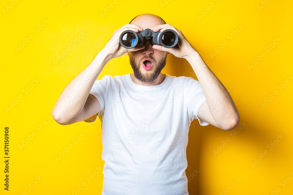 Surprised shocked bearded young man looks through binoculars on a yellow background Surprised shocked bearded young man looks through binoculars on a yellow background
